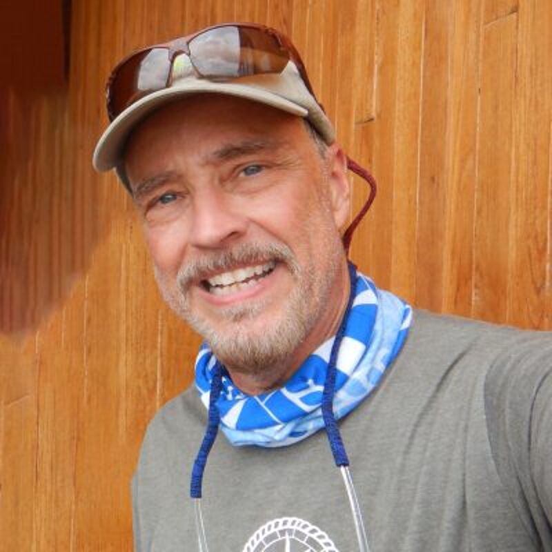 The image shows a middle-aged man with a friendly expression. He's wearing a cap, sunglasses, and a blue and white neck gaiter. He has a beard and is smiling. The background is a wooden wall. The man appears to be outdoors, possibly enjoying a sunny day.